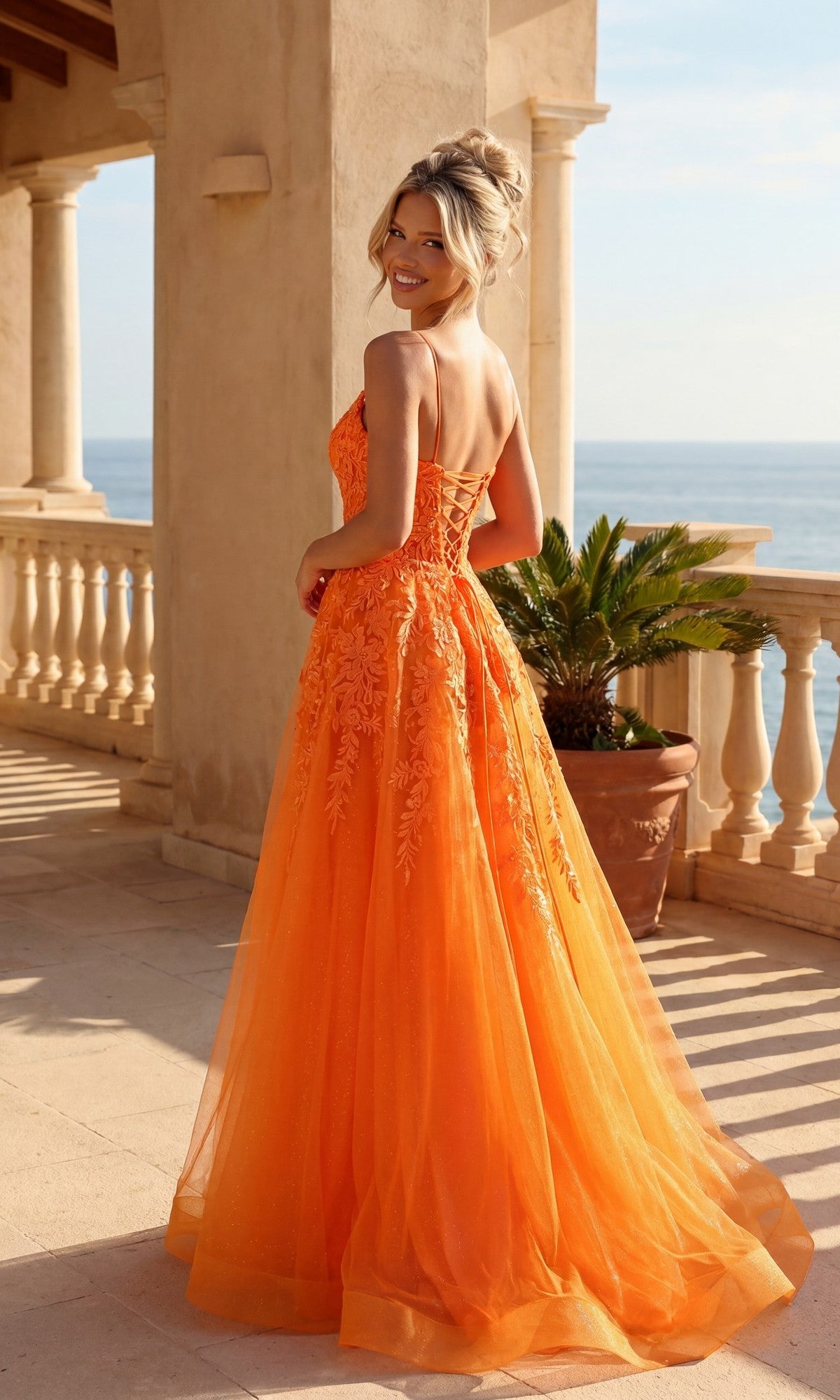 A woman in the PromGirl 250032 Glitter Tulle A-Line Prom Dress, its orange shimmer catching the sun, stands on a terrace overlooking the ocean and smiles over her shoulder.